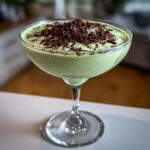 A creamy, pale green mint white chocolate martini topped generously with dark chocolate shavings in a coupe glass.
