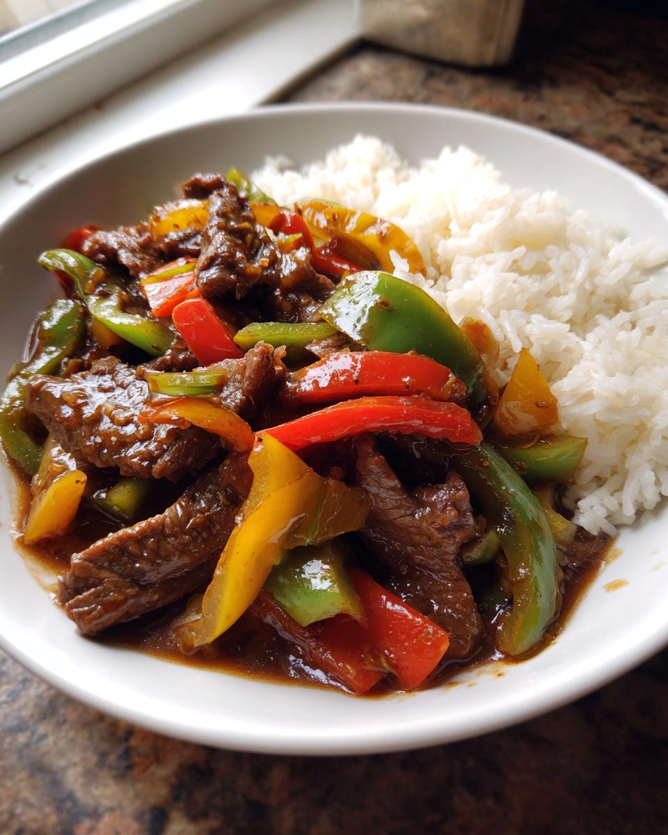 A bowl of savory crockpot Chinese pepper steak with colorful bell peppers served next to white rice.