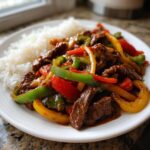 Close-up of Crockpot Chinese Pepper Steak with colorful bell peppers served next to white rice on a white plate.
