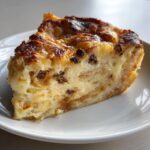 A close-up of a moist slice of croissant bread pudding with a golden-brown top and visible chocolate chips.