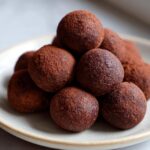 A close-up of fudgy date brownie balls stacked on a light plate, heavily dusted with cocoa powder.