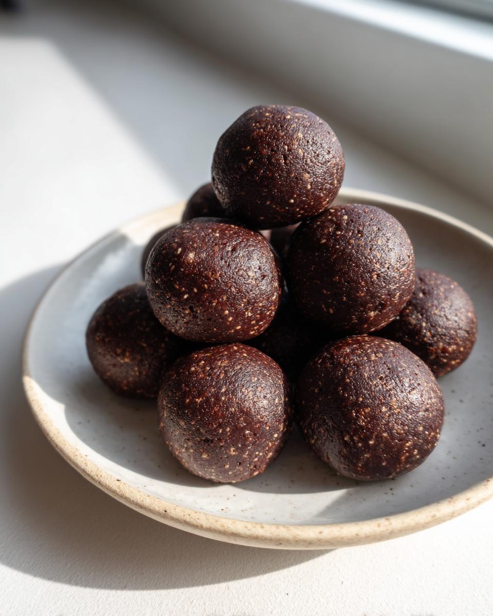 A stack of rich, dark date brownie balls piled on a speckled ceramic plate, highlighted by sunlight.