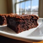 A close-up, fudgy slice of date brownies sits on a white plate, backlit by a window.
