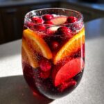 Close-up of a glass filled with bubbly Christmas sangria, loaded with orange slices, cranberries, and red apple pieces.