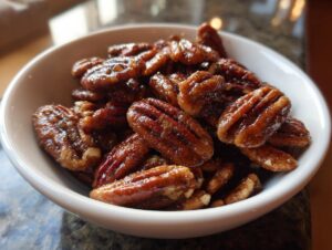 A close-up shot of glossy, freshly made candied pecans piled high in a small white bowl.