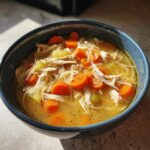 Close-up of a blue bowl filled with gluten free slow cooker chicken noodle soup, featuring shredded chicken, carrots, and celery.