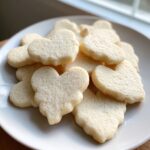 A pile of freshly baked, heart-shaped vegan sugar cookies resting on a white plate near a window.