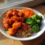 A close-up of a hot honey chicken bowl featuring glazed chicken pieces over brown rice with steamed broccoli and shredded carrots.