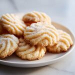 A stack of freshly baked Italian butter cookies with ridged tops, dusted with sugar, resting on a white plate.