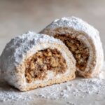 Close-up of two halves of Italian nut roll cookies, showing the rich walnut filling and heavy dusting of powdered sugar.