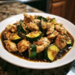 A close-up of keto chicken zucchini stir fry coated in a savory sauce and topped with sesame seeds on a white plate.