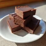 A stack of rich, dark squares of keto chocolate peanut butter fudge resting on a small white plate.