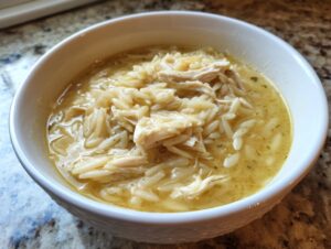 Close-up of a white bowl filled with rich lemon chicken orzo soup, featuring shredded chicken and orzo pasta.