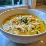Close-up of a white bowl filled with bright yellow lemon chicken orzo soup, featuring shredded chicken, orzo pasta, and carrots.
