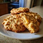 A stack of freshly baked oatmeal coconut cookies on a white plate, one cookie broken open to show the chewy texture.