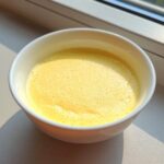 Close-up of a bright yellow, smooth steamed egg custard with tiny bubbles on top, served in a white bowl.