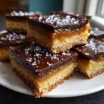 Close-up of stacked salted caramel fudge bars with a shortbread base, caramel layer, chocolate topping, and sea salt flakes.