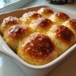 Close-up of freshly baked, golden brown soft dinner rolls glistening with a sweet glaze in a white baking dish.