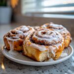 Close-up of three fluffy sourdough cinnamon rolls generously topped with white icing dripping down the sides.