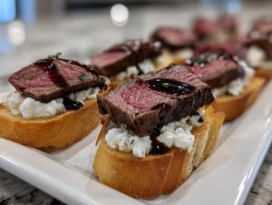 Close-up of medium-rare steak crostini topped with white cheese and balsamic glaze on a white platter.