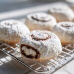 Close-up of freshly baked tasteful bites italian nut roll cookies dusted heavily with powdered sugar on a wire rack.