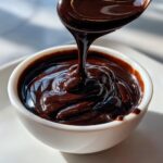 A spoonful of thick, glossy homemade chocolate syrup is being poured into a small white bowl.