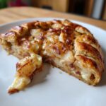 A warm slice of brie and apple galette on a white plate, showing flaky crust and melted brie cheese oozing out.