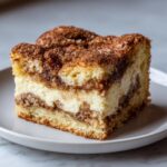 A close-up of a square slice of brown butter cream cheese snickerdoodle cobbler with layers of cake, cream cheese filling, and cinnamon topping.