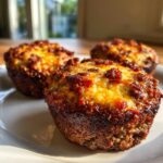 Three freshly baked carnivore pizza muffins with a crispy, browned meat base and melted, golden cheese topping on a white plate.