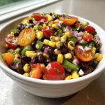 Close-up of a white bowl filled with colorful cowboy caviar recipe ingredients: black beans, corn, cherry tomatoes, and red onion.
